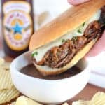 Close up of french dip sandwich being dipped into a bowl of au jus.