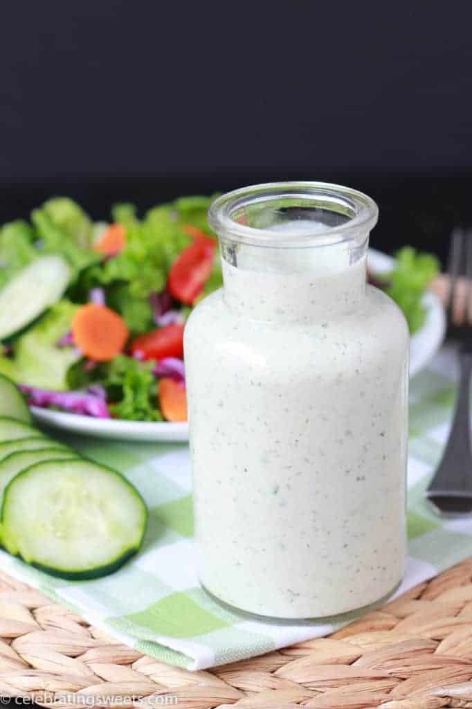 Salad dressing in a glass container with a salad in the background. 