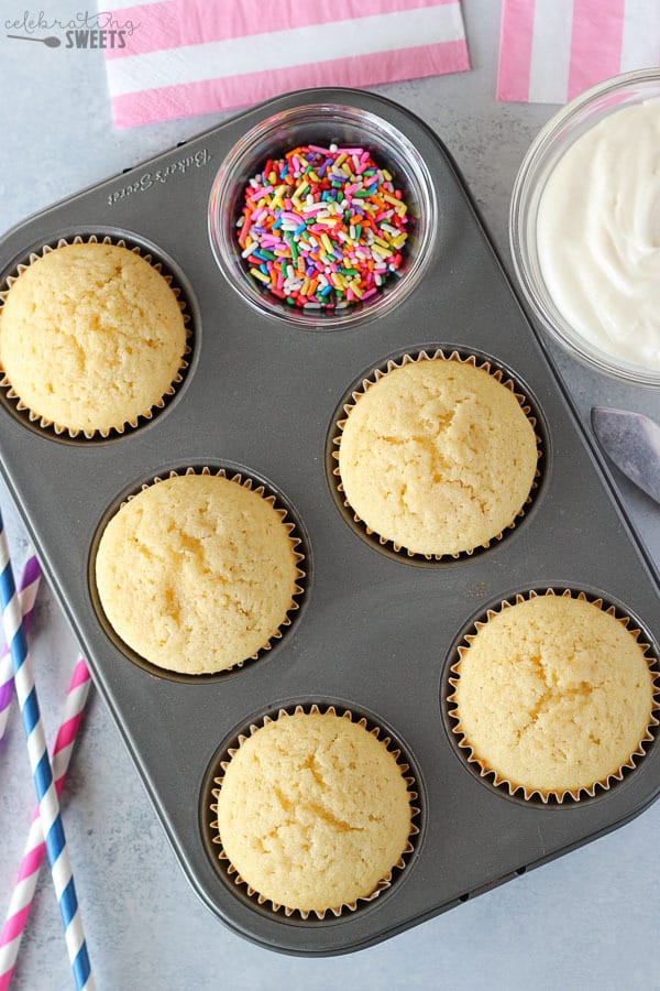 Vanilla cupcakes in a pan with a bowl of frosting and a bowl of sprinkles.