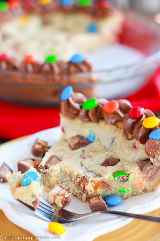 Slice of cookie cake filled with candy bars and topped with chocolate frosting.