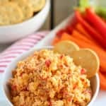 White bowl filled with pimento cheese garnished with crackers and vegetables.