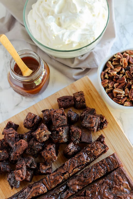 Chopped brownies on a cutting board. Bowls of caramel sauce, chopped pecans, and whipped cream.