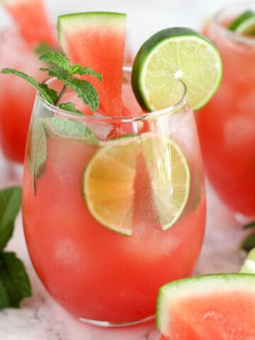 Pink Watermelon Mojitos in a glasses garnished with mint, lime, and watermelon.