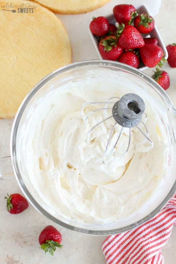 Bowl of whipped cream with layers of cake and strawberries in the background.