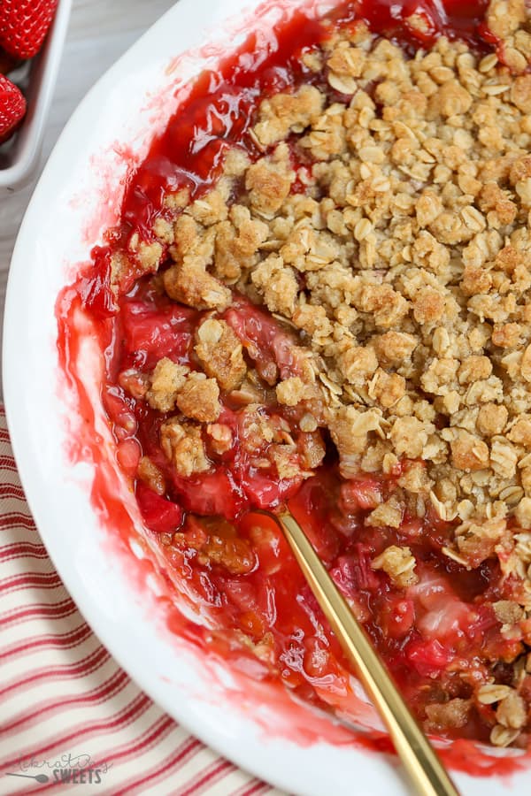 Strawberry rhubarb crisp in a white baking dish with a gold spoon.
