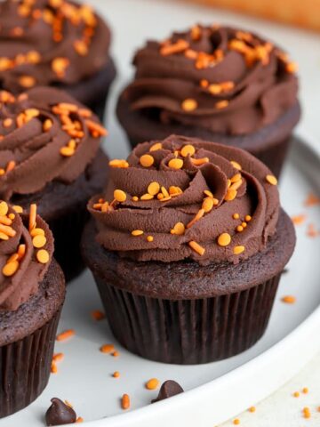 Chocolate cupcakes with chocolate frosting and orange sprinkles on a white plate.