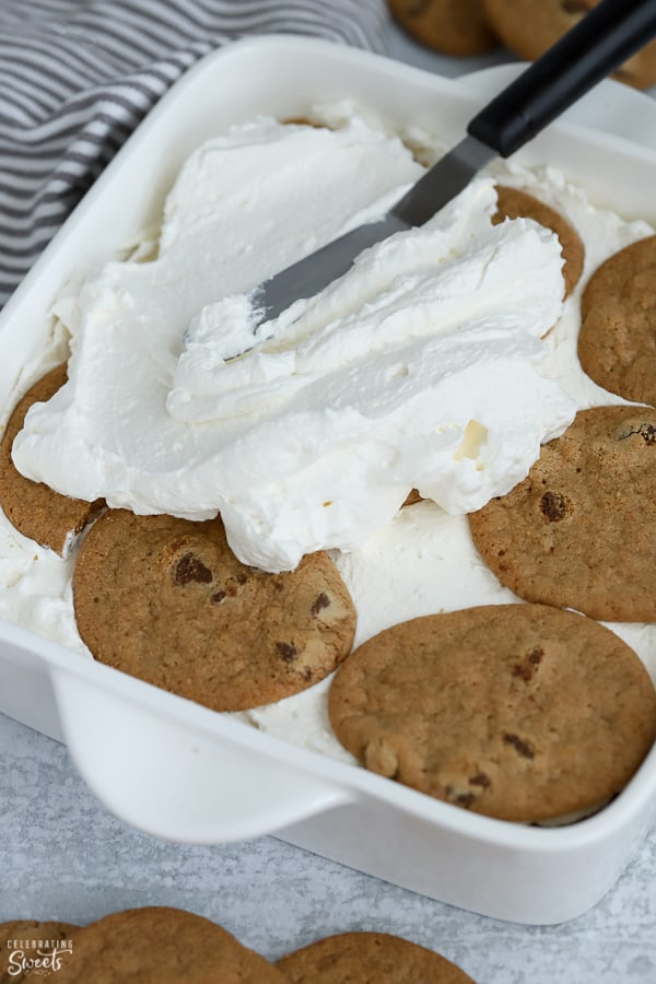 Whipped cream being spread over chocolate chip cookies in a white baking dish.