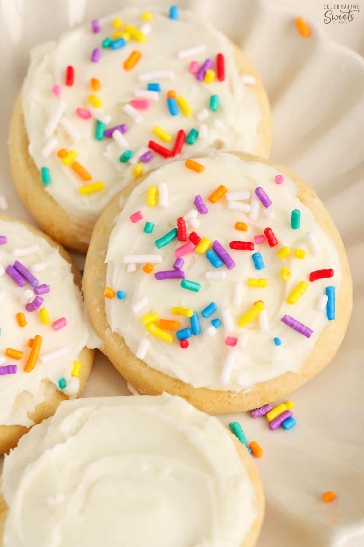 Frosted sugar cookies topped with rainbow sprinkles on a white plate.