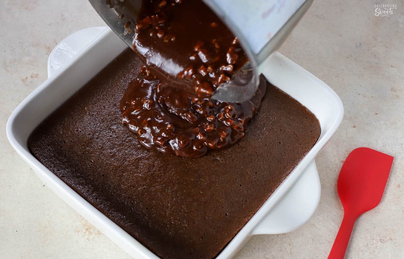 Chocolate icing being poured over chocolate cake in a white pan.