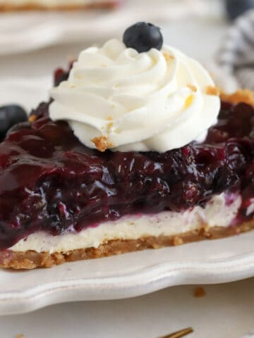 Slice of Blueberry Pie on a white plate topped with whipped cream.