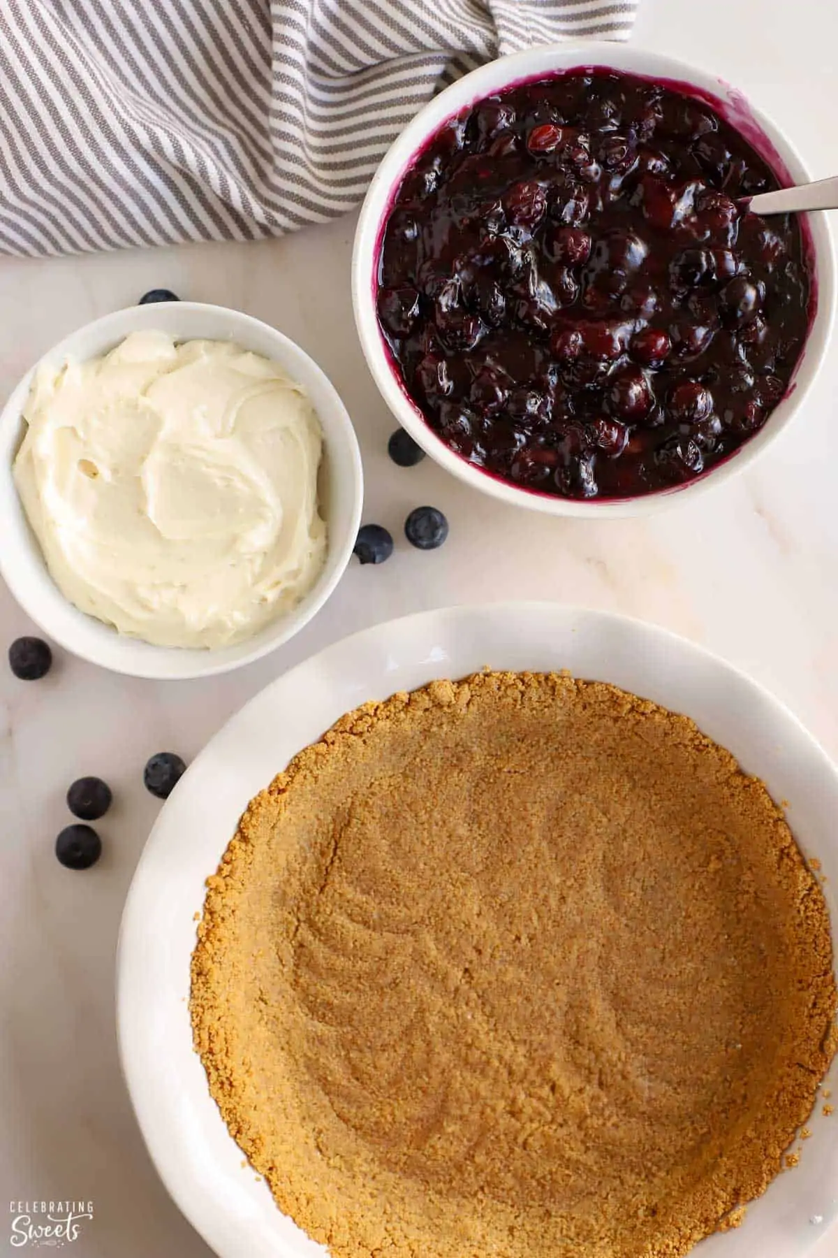 Graham cracker crust in a white pie plate, blueberry pie filling in a white bowl, and cream cheese in a white bowl.