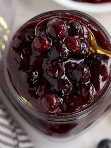 Blueberry Pie Filling in a glass jar with a gold spoon.