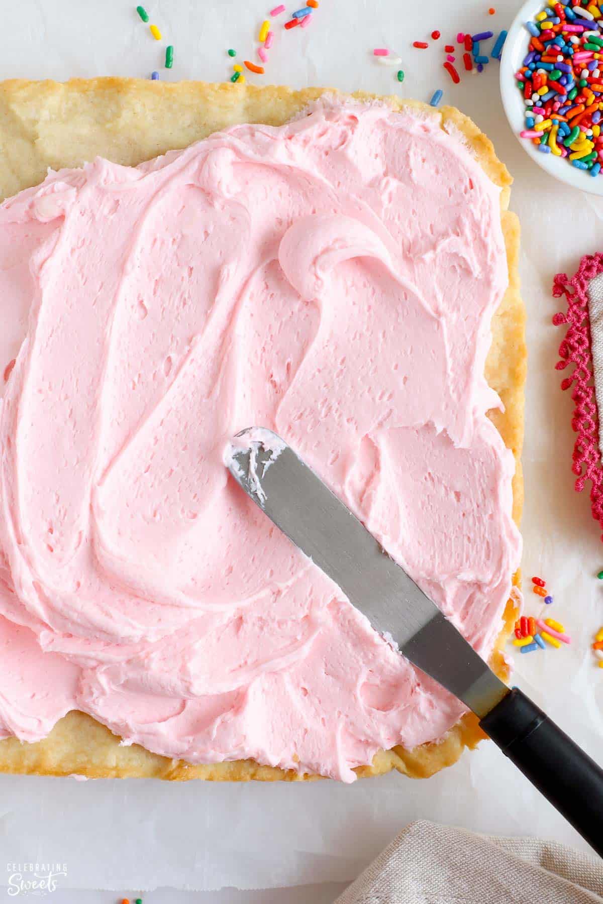 Pink frosting spread onto sugar cookie bars surrounded by sprinkles.