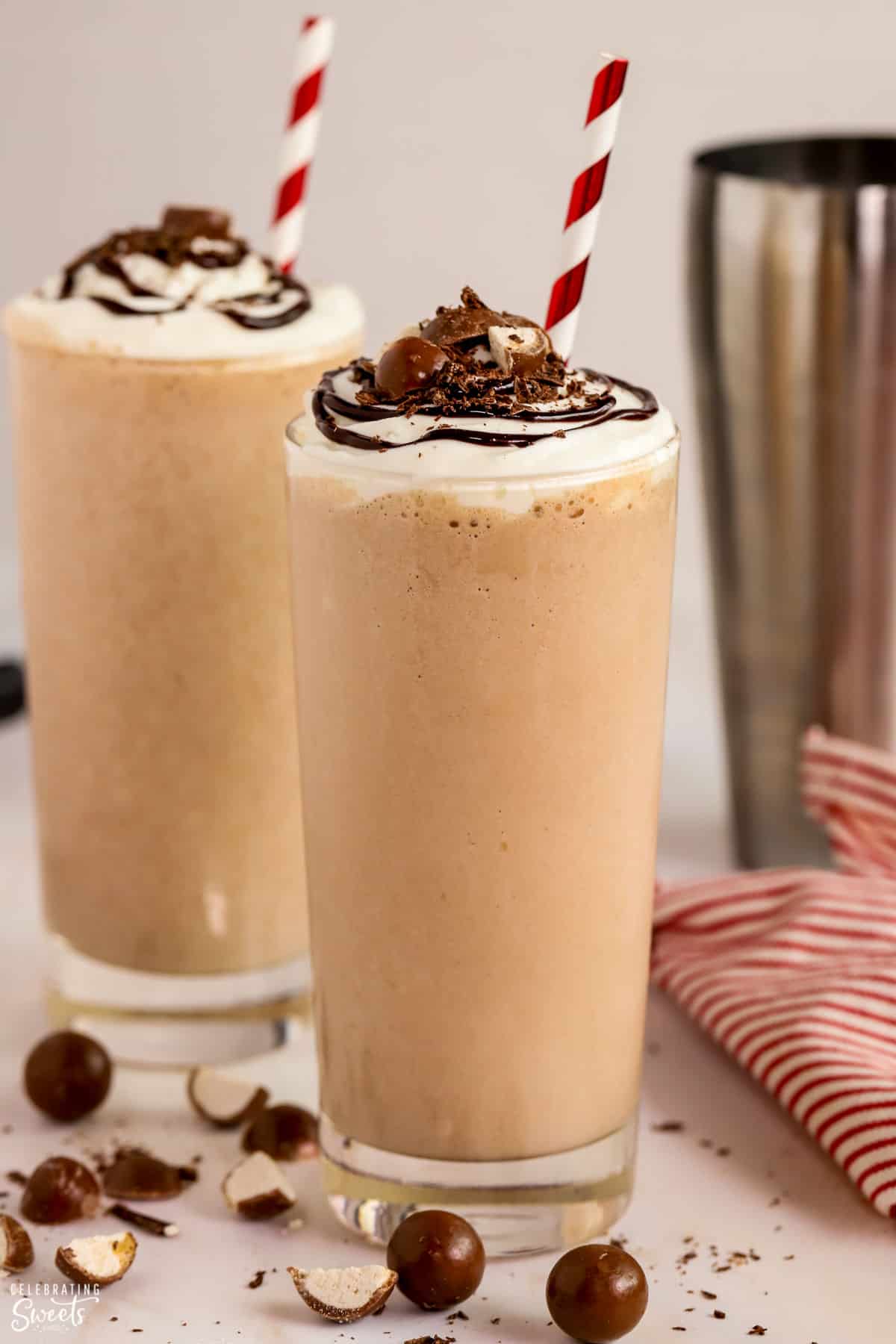 Two glasses filled with chocolate malt topped with whipped cream, chocolate sauce, and chopped malted milk balls.