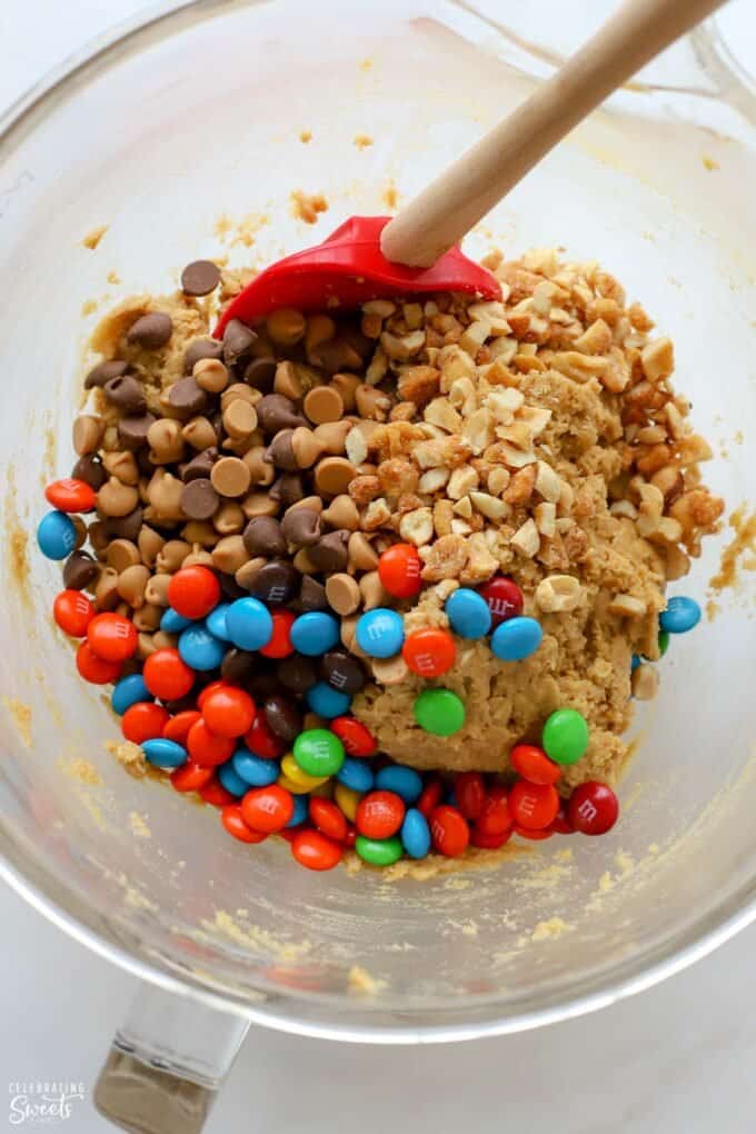 Cookie dough in a glass mixing bowl topped with chocolate chips, peanut butter chips, M&M's, and peanuts.