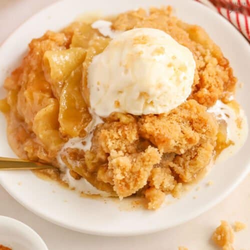Apple crumble on a white plate topped with a scoop of melty vanilla ice cream.