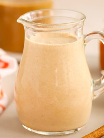 Pumpkin spice creamer in a glass carafe next to cinnamon sticks.