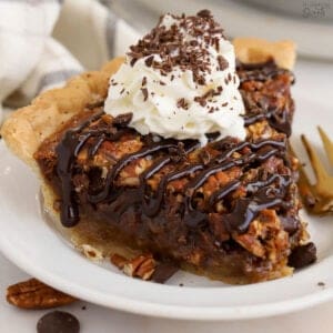 Slice of chocolate pecan pie on a white plate topped with whipped cream and chocolate sauce.