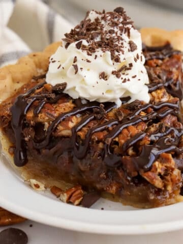 Slice of chocolate pecan pie on a white plate topped with whipped cream and chocolate sauce.