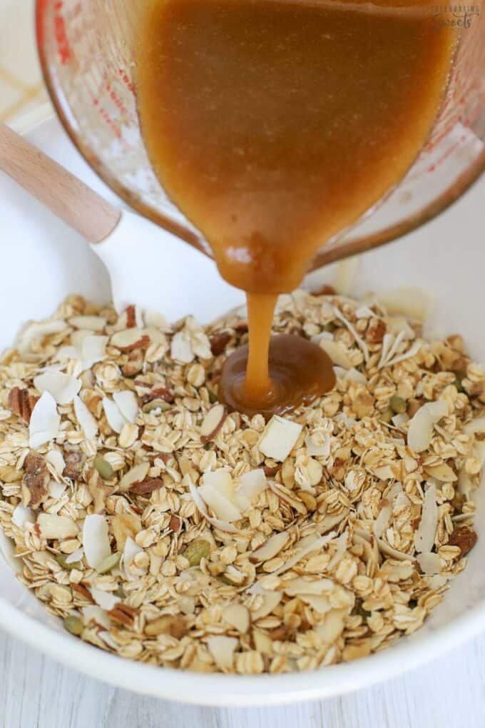 Maple syrup mixture being poured into a white bowl filled with oats and nuts.