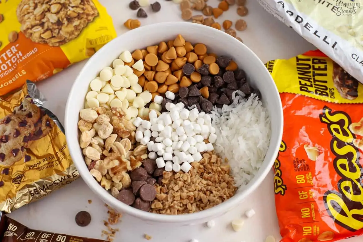 Chocolate chips, butterscotch chips, marshmallows, nuts, toffee chips, coconut in a white bowl.