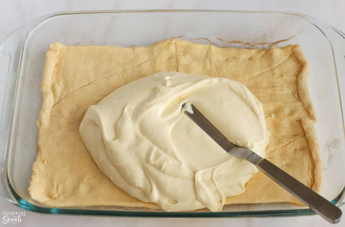 Cream cheese being spread on crescent roll dough in a glass pan.