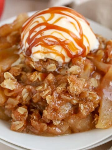 Apple crisp on a white plate topped with vanilla ice cream and caramel sauce.