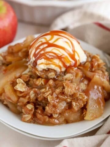 Apple crisp on a white plate topped with vanilla ice cream and caramel sauce.