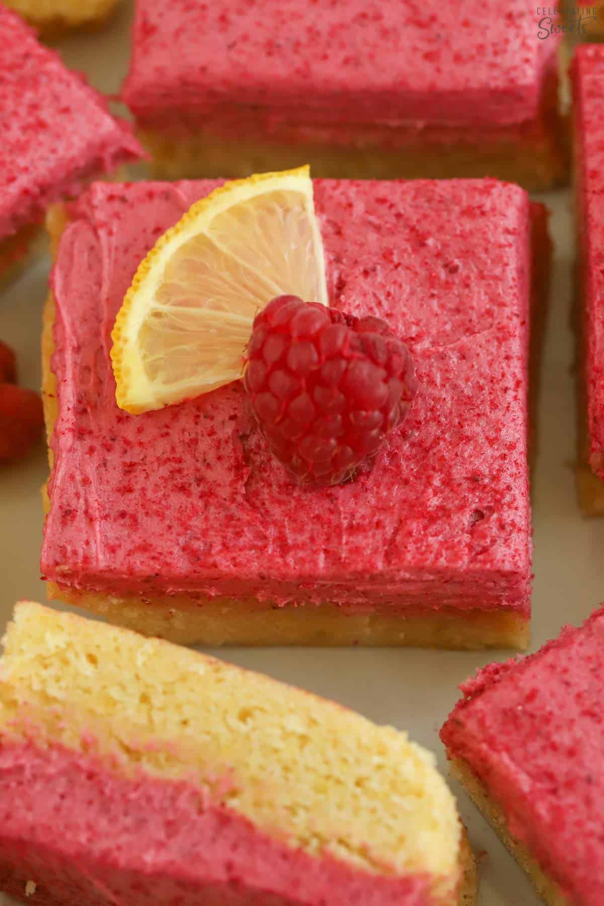 Lemon bar topped with pink frosting and garnished with a lemon slice and raspberry.