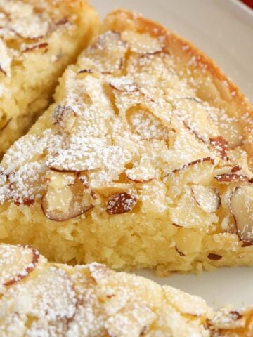 Slice of almond cake on a grey plate topped with sliced almonds and powdered sugar,