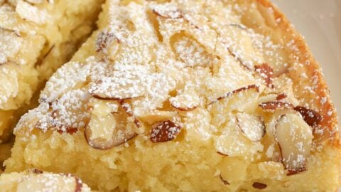 Slice of almond cake on a grey plate topped with sliced almonds and powdered sugar,