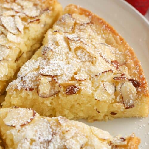 Slice of almond cake on a grey plate topped with sliced almonds and powdered sugar,