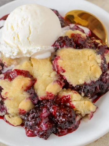 Blackberry cobbler topped with vanilla ice cream on a white plate with a gold spoon.