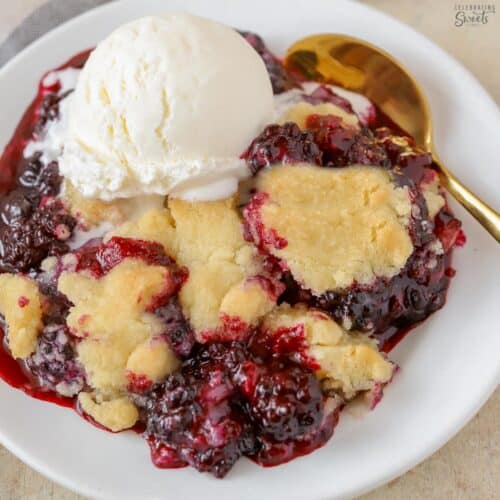 Blackberry cobbler topped with vanilla ice cream on a white plate with a gold spoon.