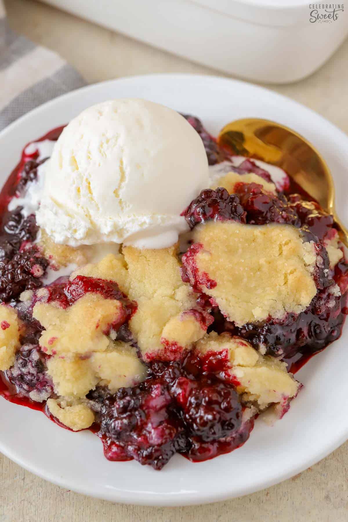 Blackberry cobbler topped with vanilla ice cream on a white plate with a gold spoon.