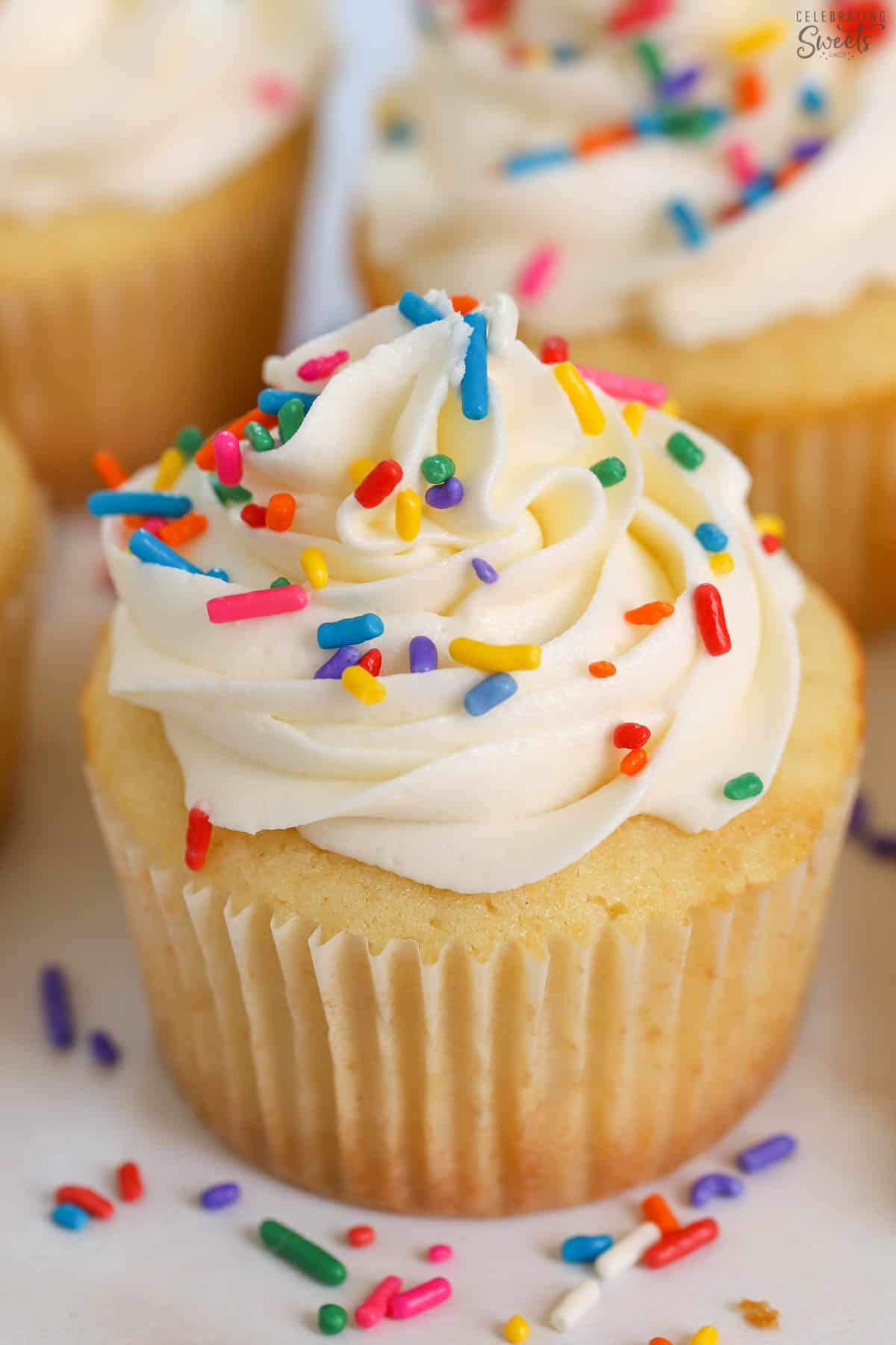 Closeup of a vanilla cupcake topped with white frosting and rainbow sprinkles.