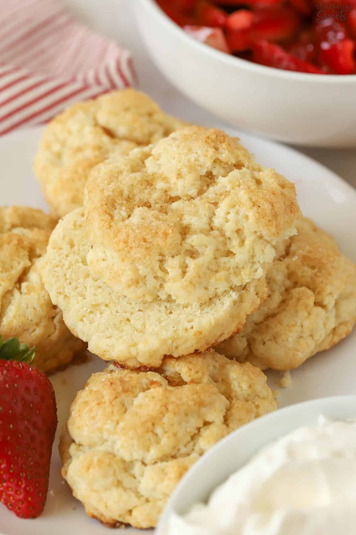 Strawberry shortcake biscuits on a white plate next to strawberries and whipped cream.