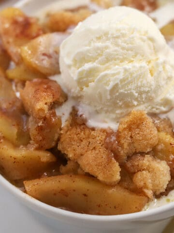 Apple cobbler in a white bowl topped with vanilla ice cream.