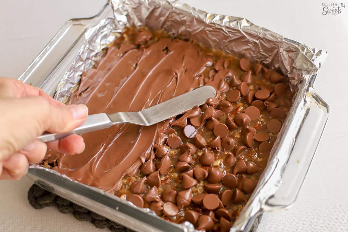 Melted chocolate being spread on caramel bars in a glass pan.