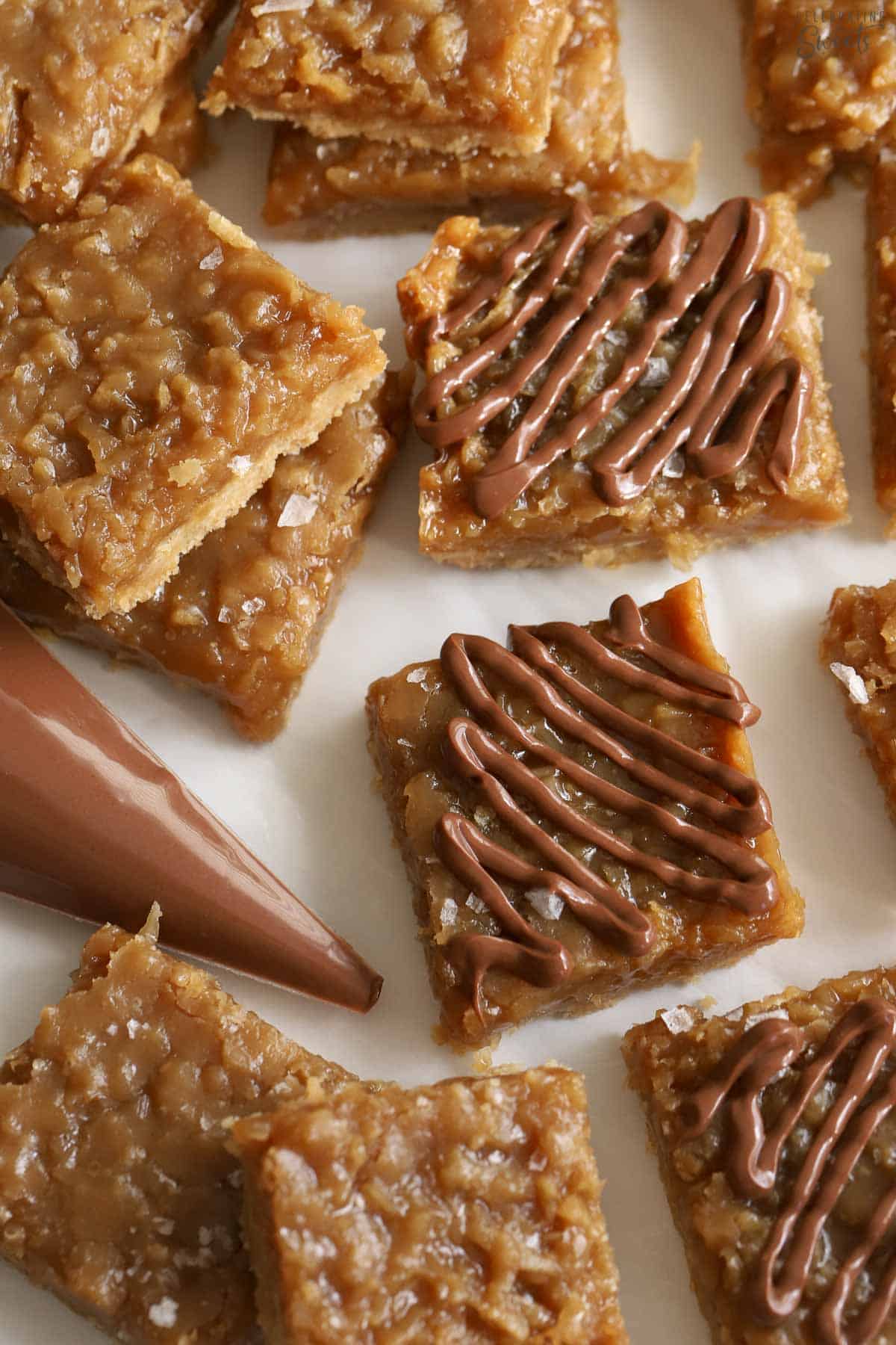 Caramel coconut bars on a piece of parchment paper next to a piping bag filled with melted chocolate.