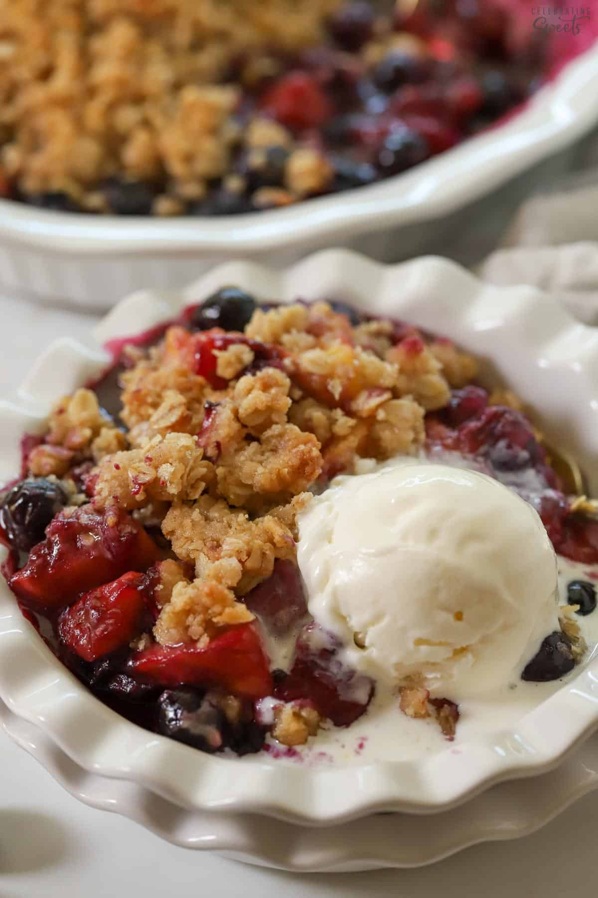 Blueberry peach crisp in a white bowl with a scoop of vanilla ice cream.