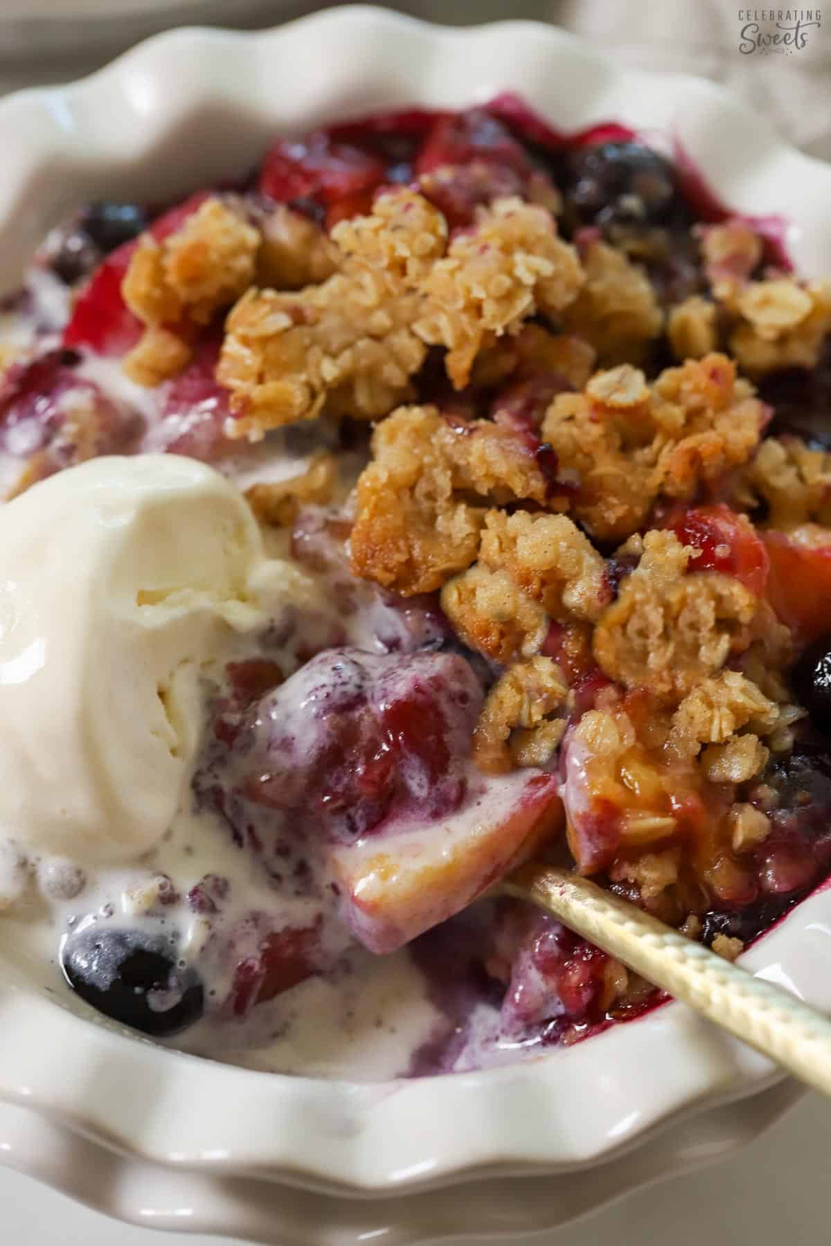 Closeup of a blueberry peach crisp in a white bowl topped with vanilla ice cream.
