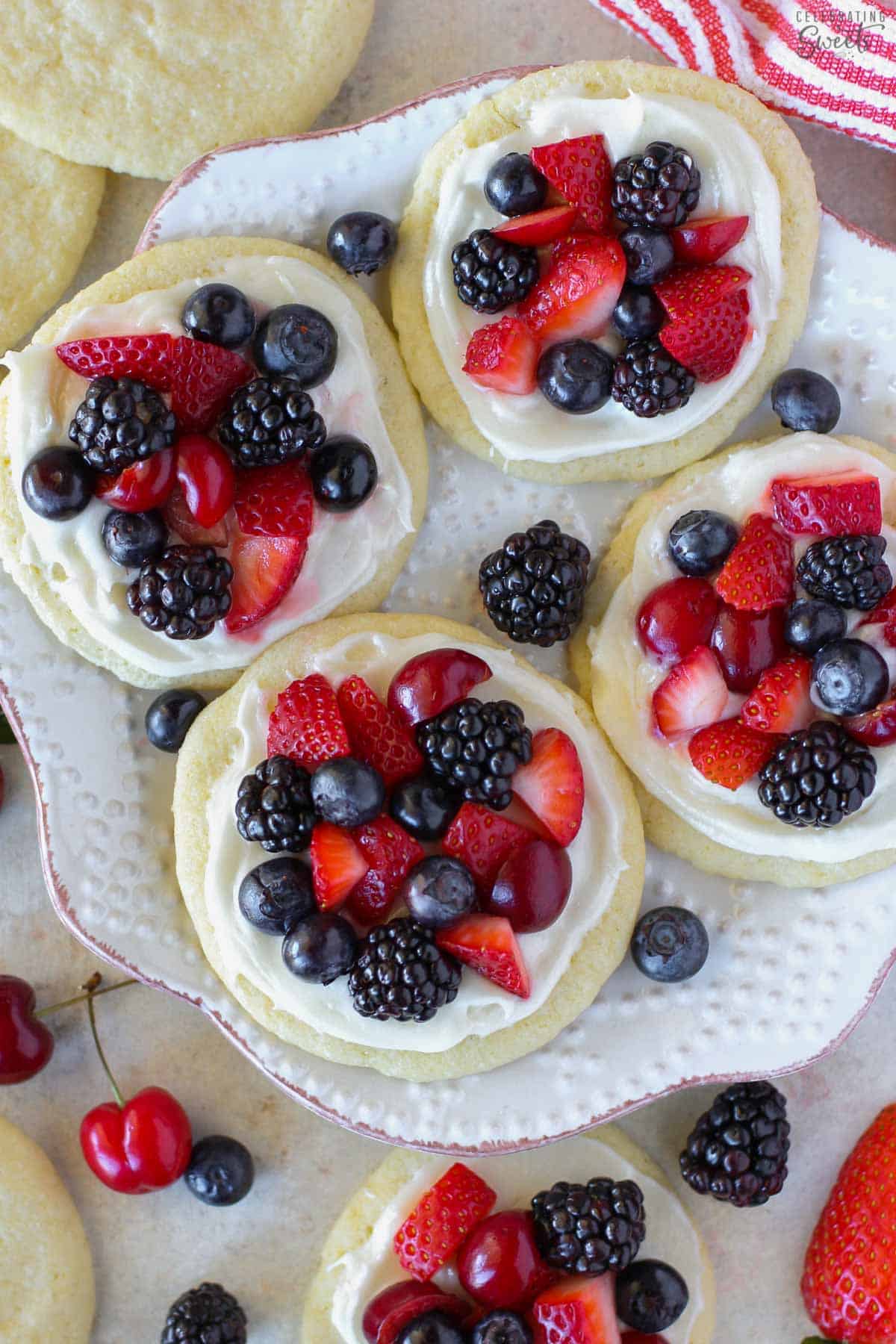 Four sugar cookie fruit tarts topped with berries on a white plate surrounded by berries and cherries.