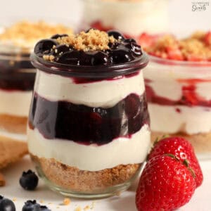 Cheesecake parfait with blueberry sauce layered in a glass jar next to fresh berries.