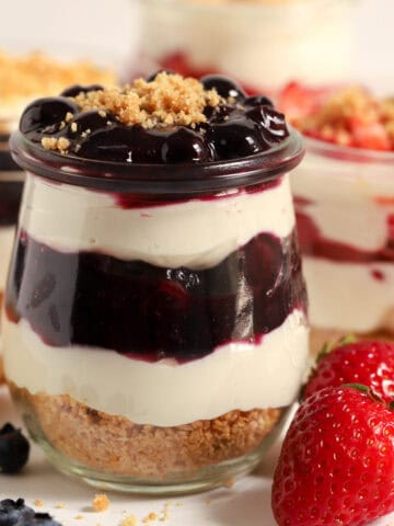 Cheesecake parfait with blueberry sauce layered in a glass jar next to fresh berries.