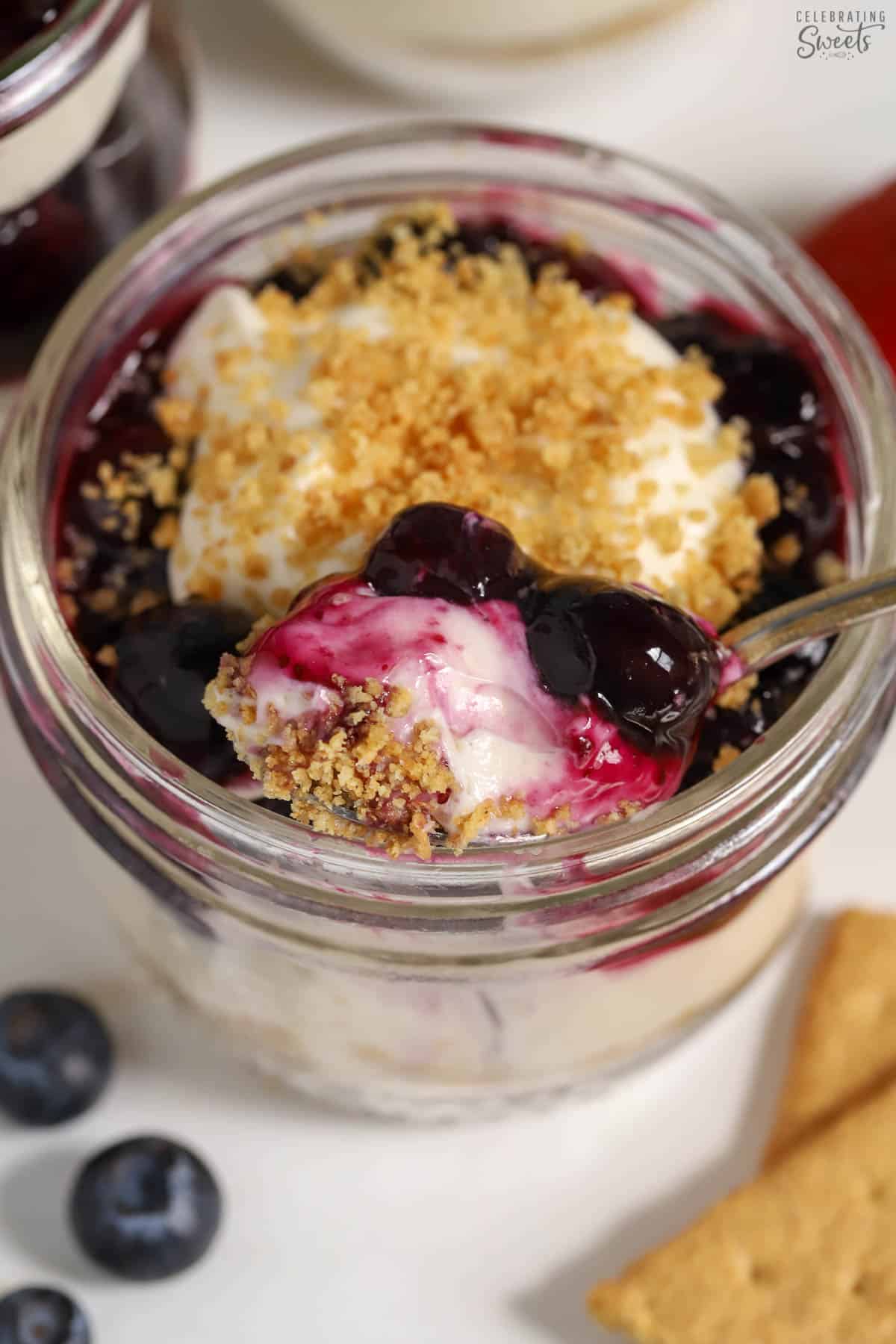 Cheesecake parfait in a glass jar with blueberry sauce and a gold and silver spoon.