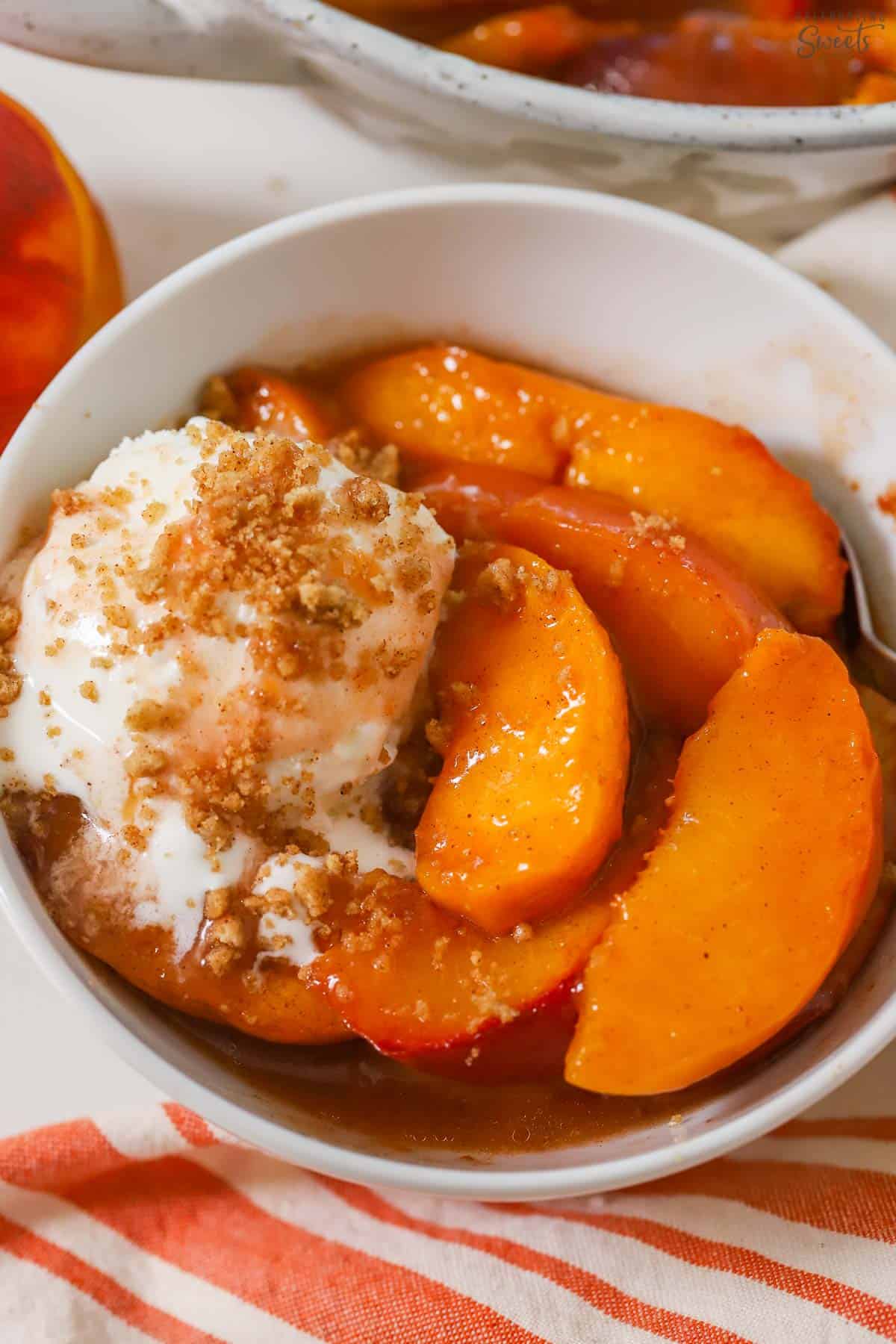 Baked peaches in a white bowl topped with vanilla ice cream and cookie crumbs.