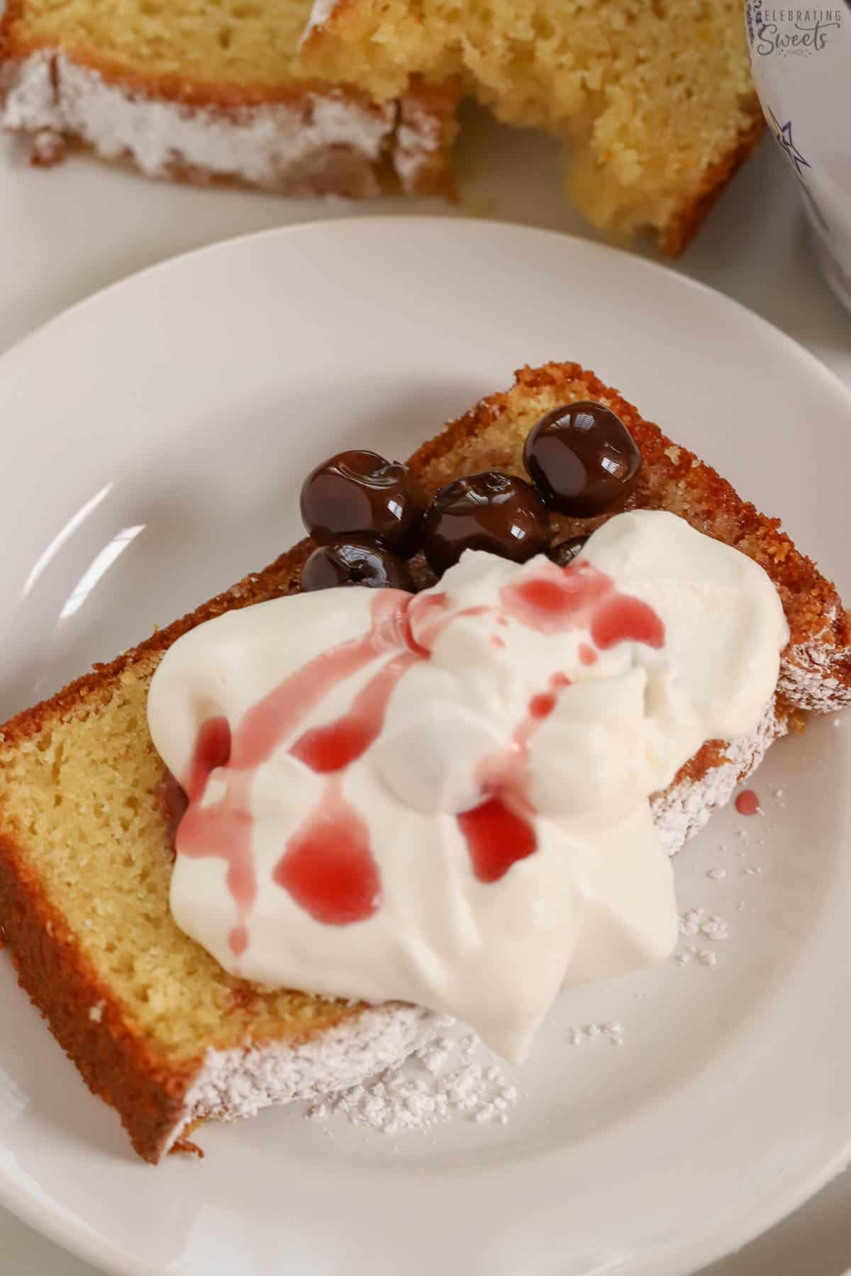 A slice of yogurt cake on a white plate topped with cherries and whipped cream.