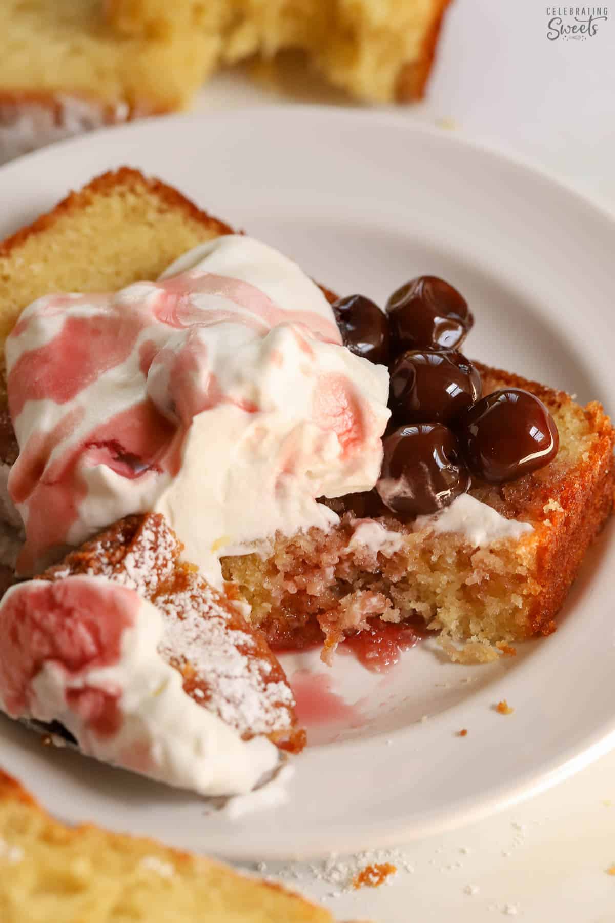 Slice of yogurt cake on a white plate topped with whipped cream and cherries.