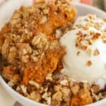 Closeup of a pumpkin crisp in a white bowl topped with vanilla ice cream and chopped pecans.